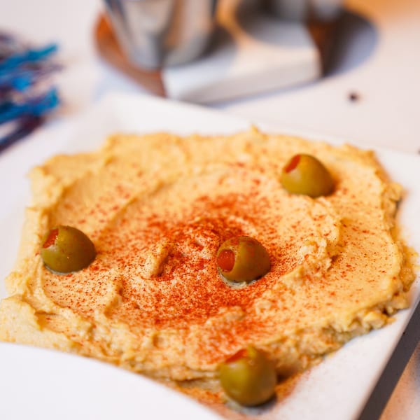 Creamy hummus topped with red paprika and green olives, served on a white plate.