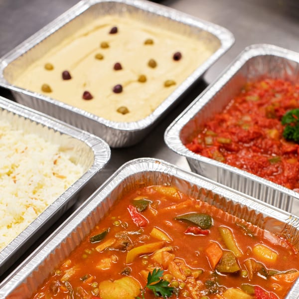 Four aluminum trays filled with various Mediterranean dishes, including hummus, rice, and stews.