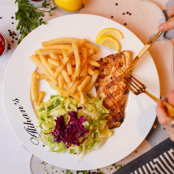 A person cuts into a grilled fish fillet with fries and salad on an Alihan's branded plate.